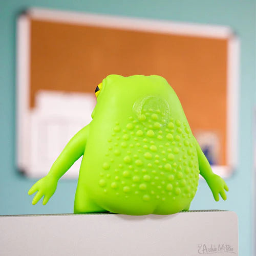 Bright green splat frog toy with warty back and tiny bum sitting on edge of a desk or monitor in a colorful room