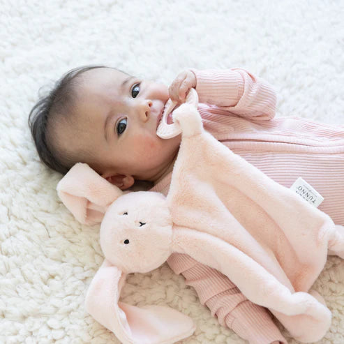 Infant lying on soft blanket holding and chewing on a soft plush bunny teether buddy toy