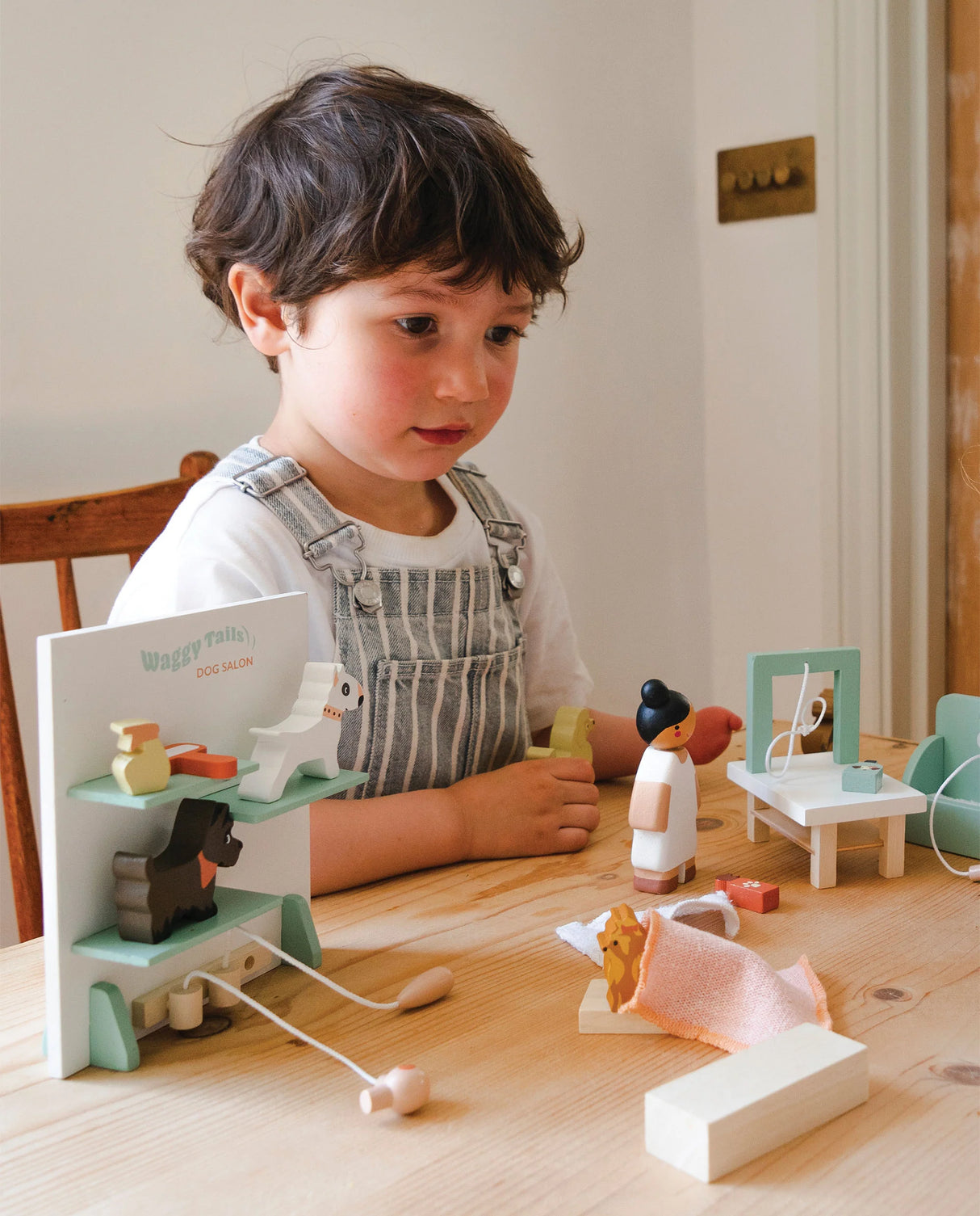 Child playing with Waggy Tails Dog Salon wooden grooming set featuring dog figures and accessories on a table.