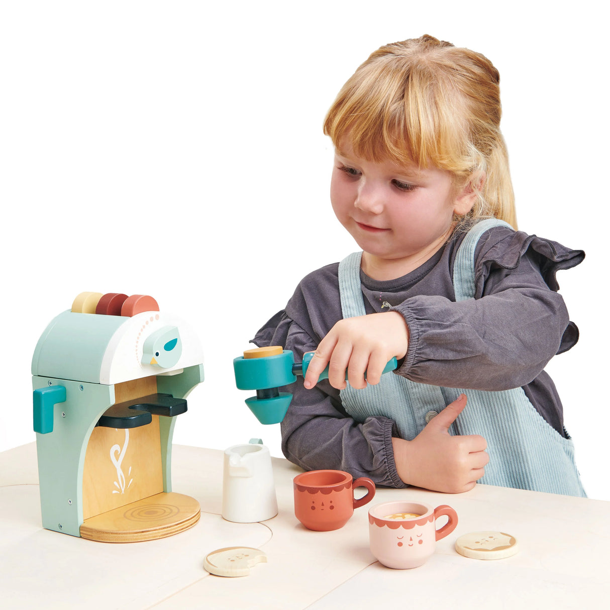 Child playing with Babyccino Maker set including coffee cups, pods, milk jug, and biscuits in soft mellow colors.