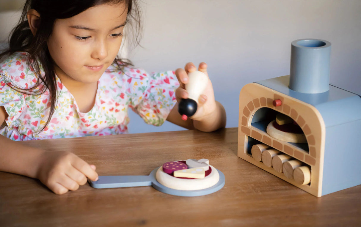 Child playing with Make Me a Pizza! toy pizza oven, using pretend toppings and pizza slider on a wooden table.