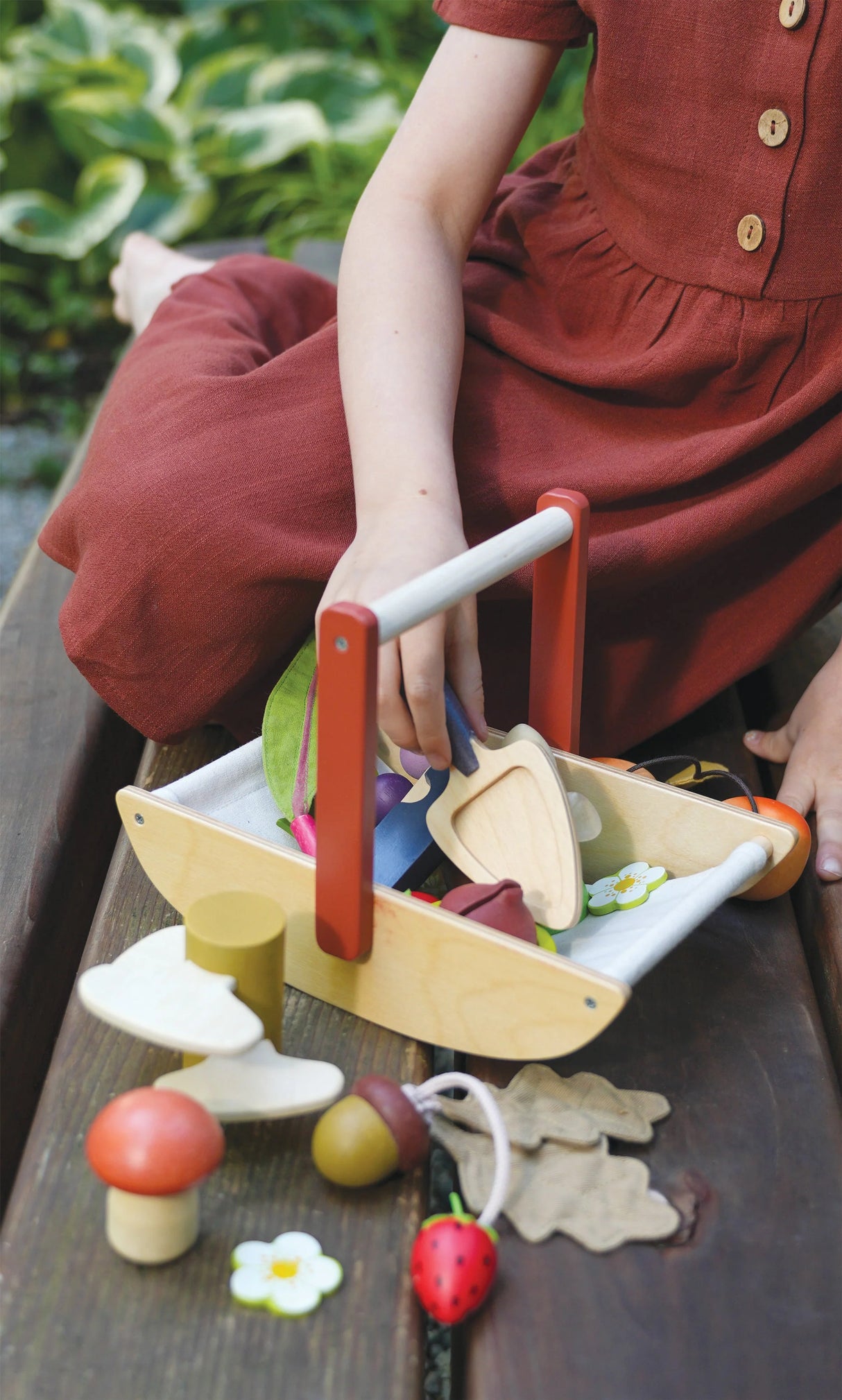 Child playing outdoors with a Wild Wood Foraging Trug filled with fabric plants, mushrooms, and flowers.