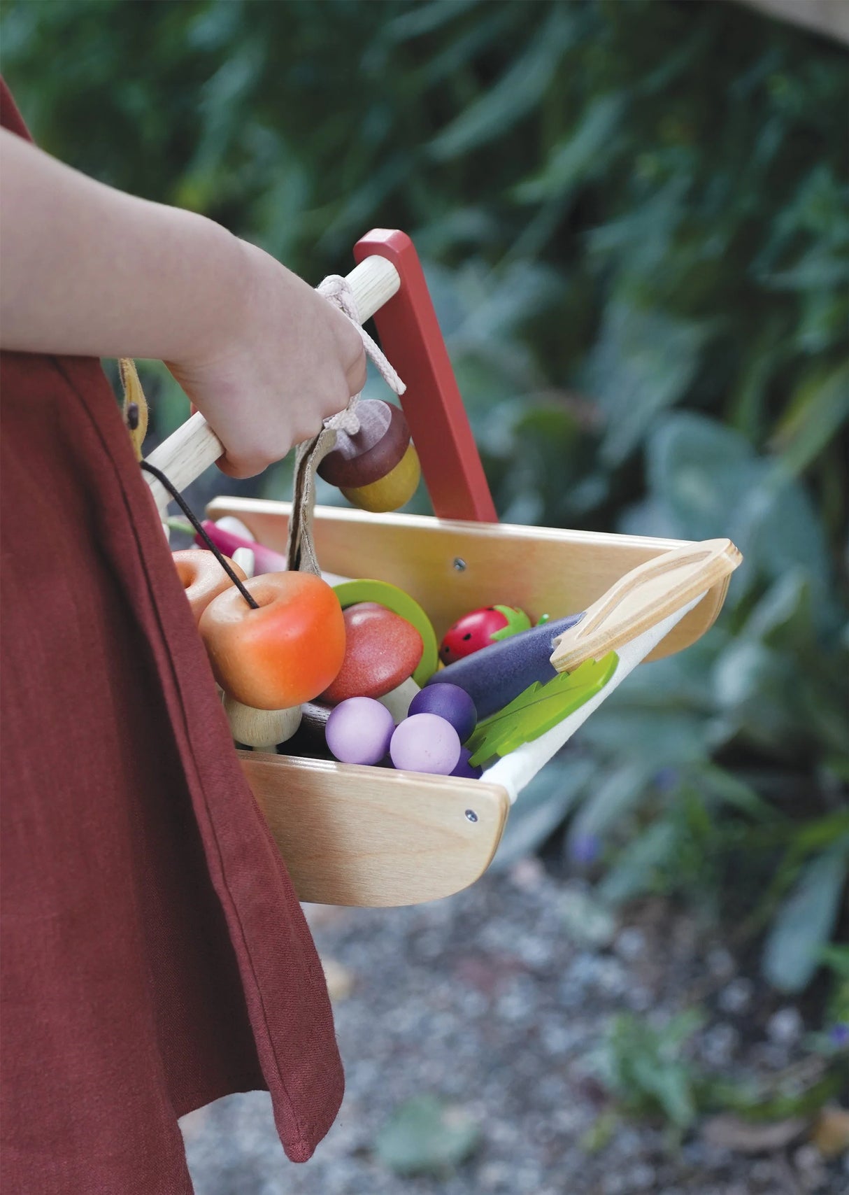 Child holding Wild Wood Foraging Trug filled with fabric fruits, vegetables, and flowers in a garden setting.