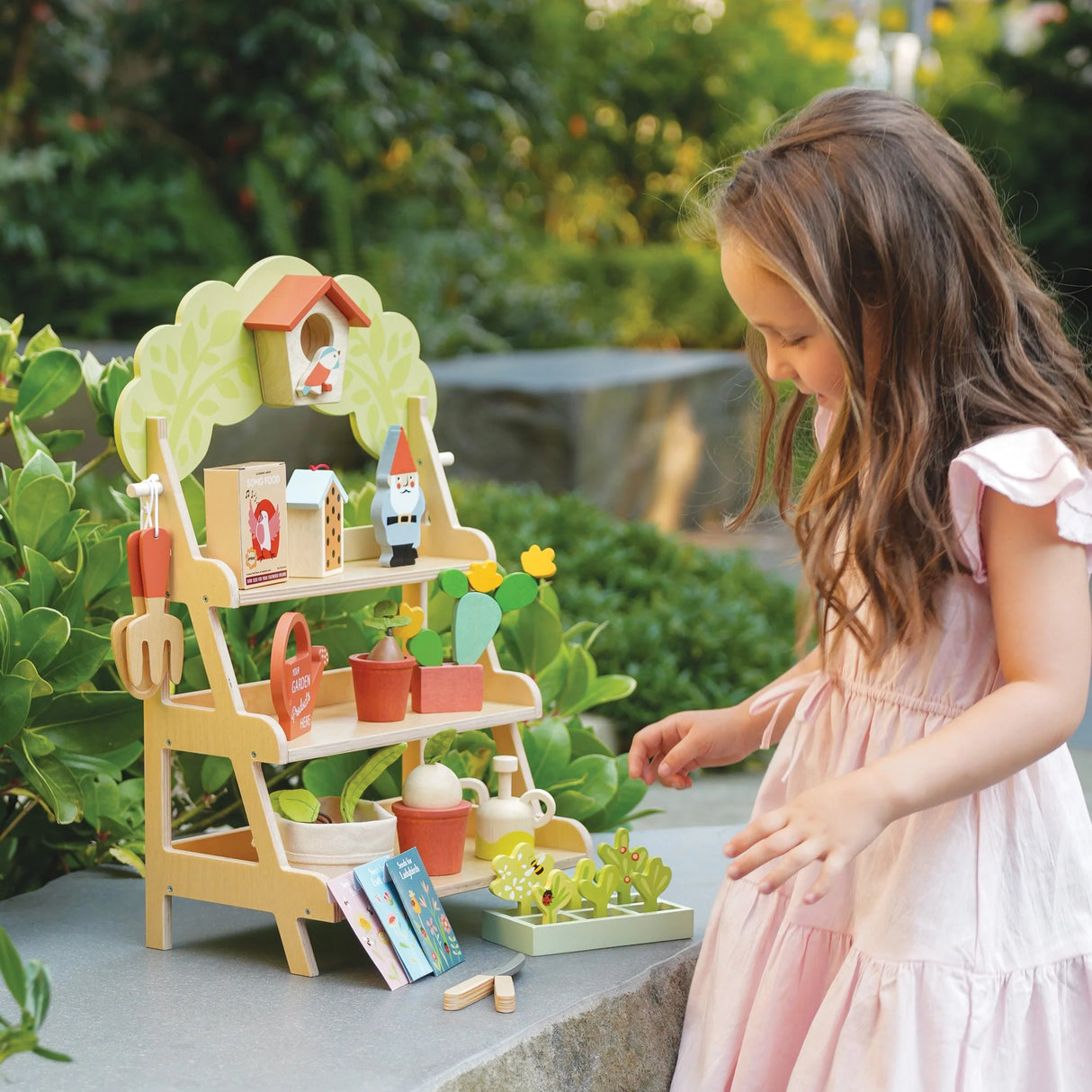 Garden Centre wooden stand with garden tools, plants, birdhouse, and girl playing outdoors in pink dress.