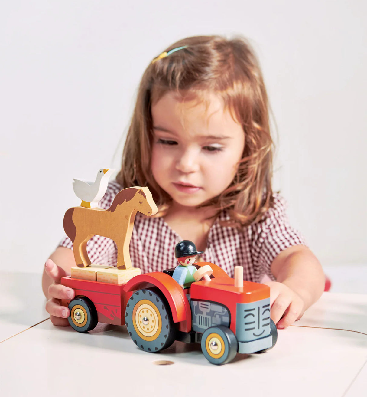 Child playing with a bright red wooden Farmyard Tractor toy with driver, trailer, horse, and goose.
