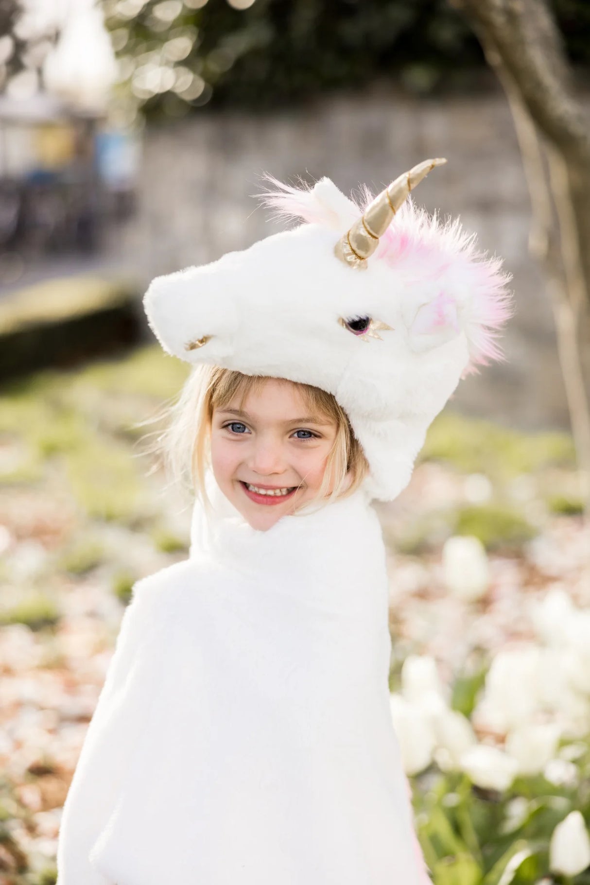 Girl smiling outdoors wearing the Unicorn Cuddle Cape Size 4-6 with a fluffy unicorn hood and gold horn.