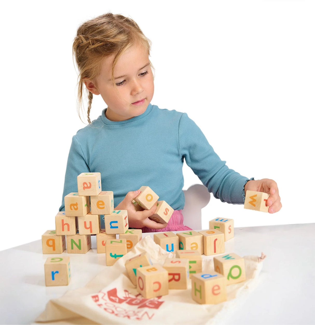 Child playing with colorful wooden alphabet blocks stacking and spelling on a white table with a drawstring bag nearby