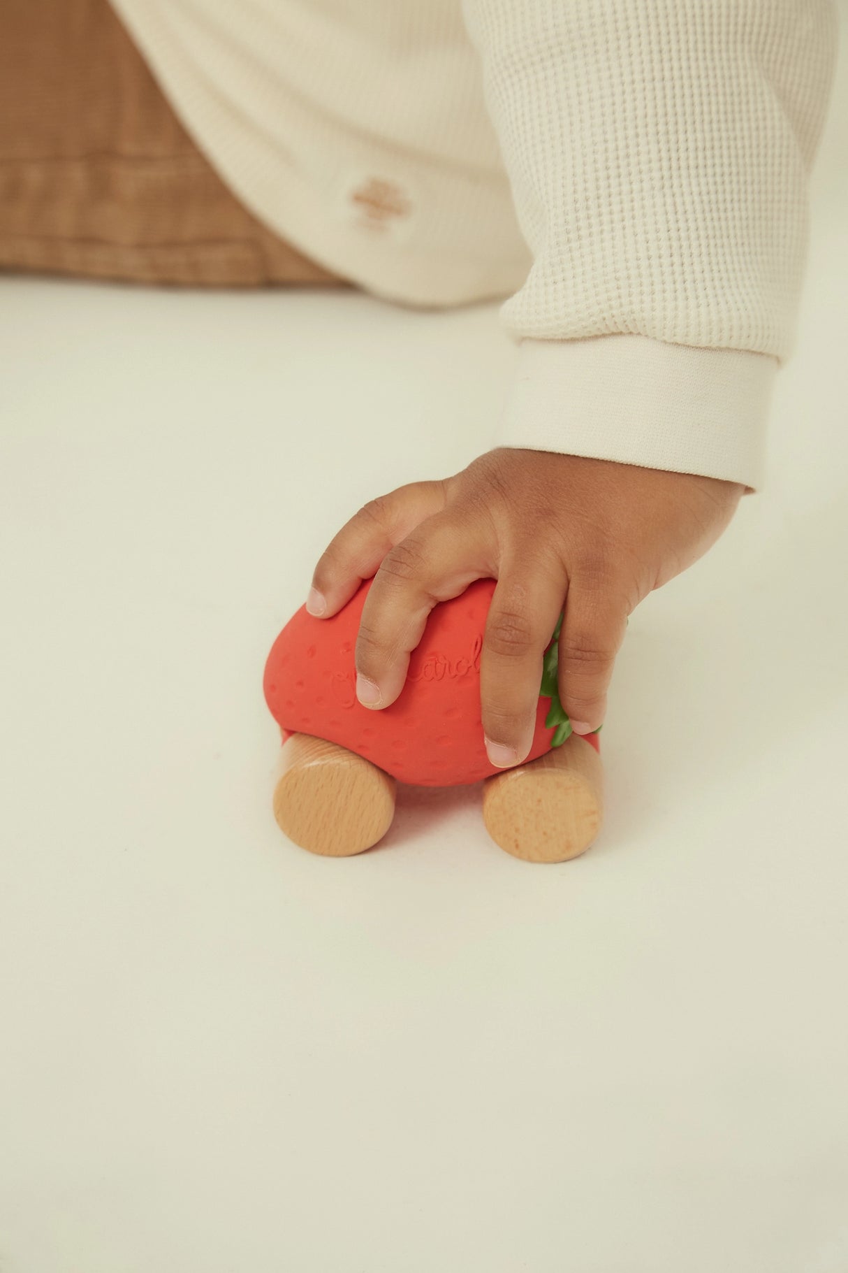 Child’s hand playing with a red strawberry-shaped baby car toy made of natural rubber and wood on white surface