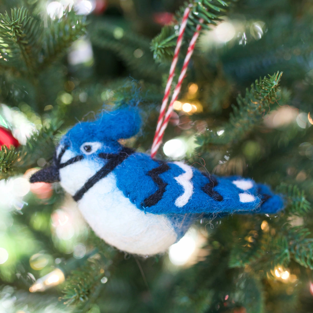 Handmade felt blue jay bird ornament hanging on a Christmas tree with green pine needles and lights background