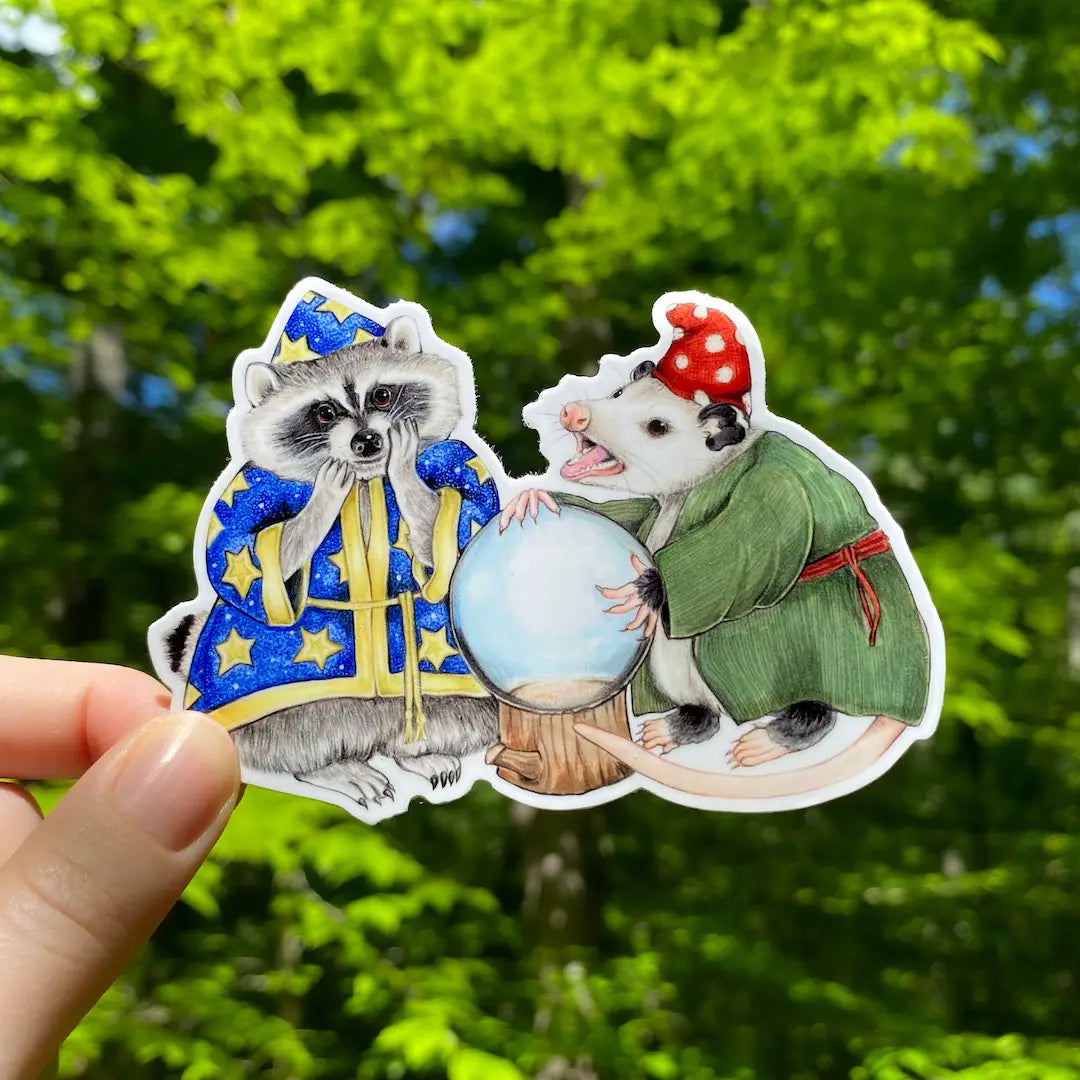 Opossum & Raccoon Fortuneteller Sticker showing a raccoon wizard and opossum with a crystal ball outdoors