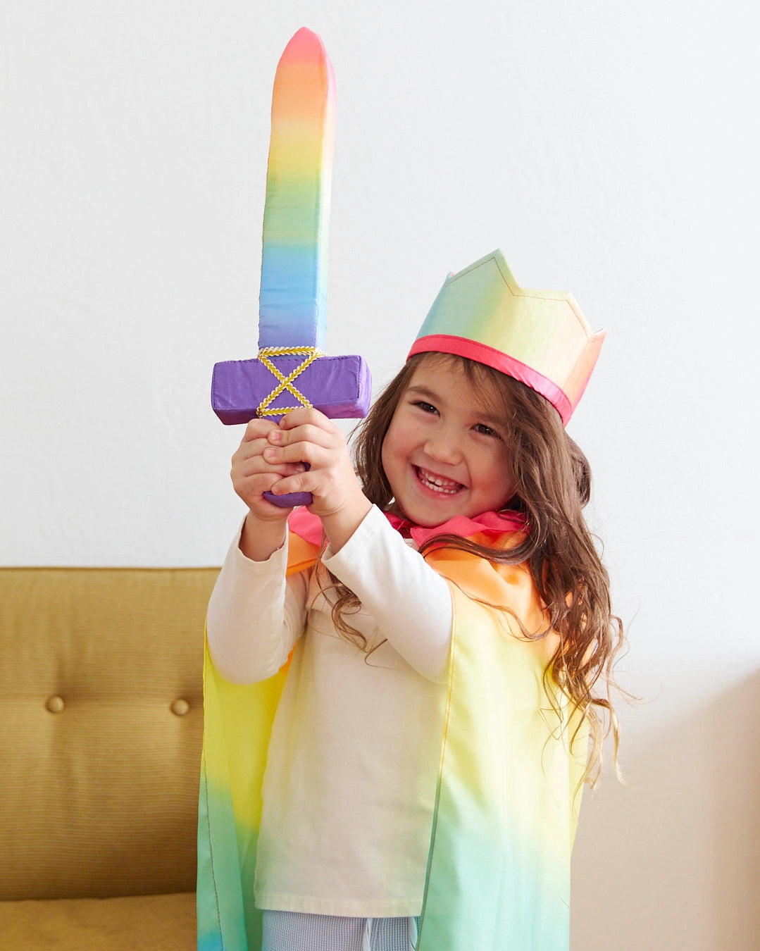 Smiling child wearing a rainbow cape and crown holding a colorful fabric sword upright indoors