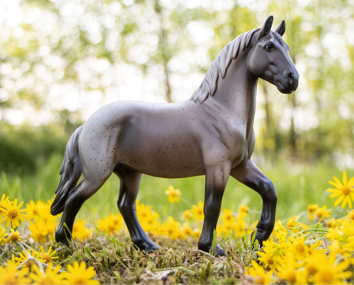 Blue Roan Brabant model horse standing among yellow flowers in a natural outdoor setting, detailed and lifelike.