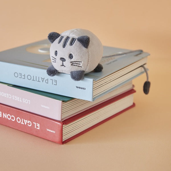 Soft fluffy grey cat shaped bookmark resting on a stack of colorful books against a beige background