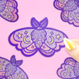 Moon Moth Glitter Sticker showing a purple glittery moth with stars and moons on a pink background.
