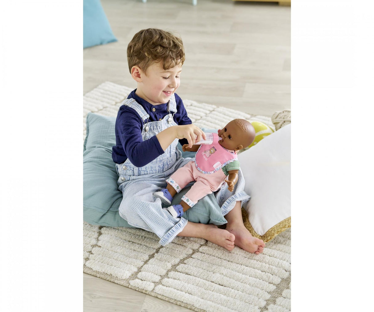 Toddler smiling and playing nurturing game by feeding a doll with a magic bottle while sitting on cushions.