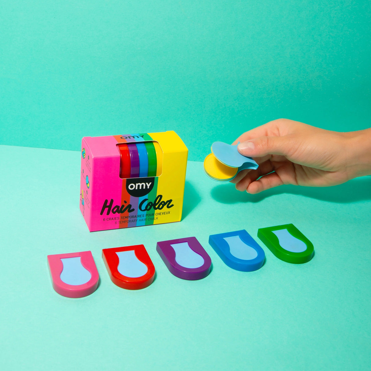 Hand holding blue hair chalk applicator with pink, red, purple, blue, and green chalks displayed next to colorful packaging box.