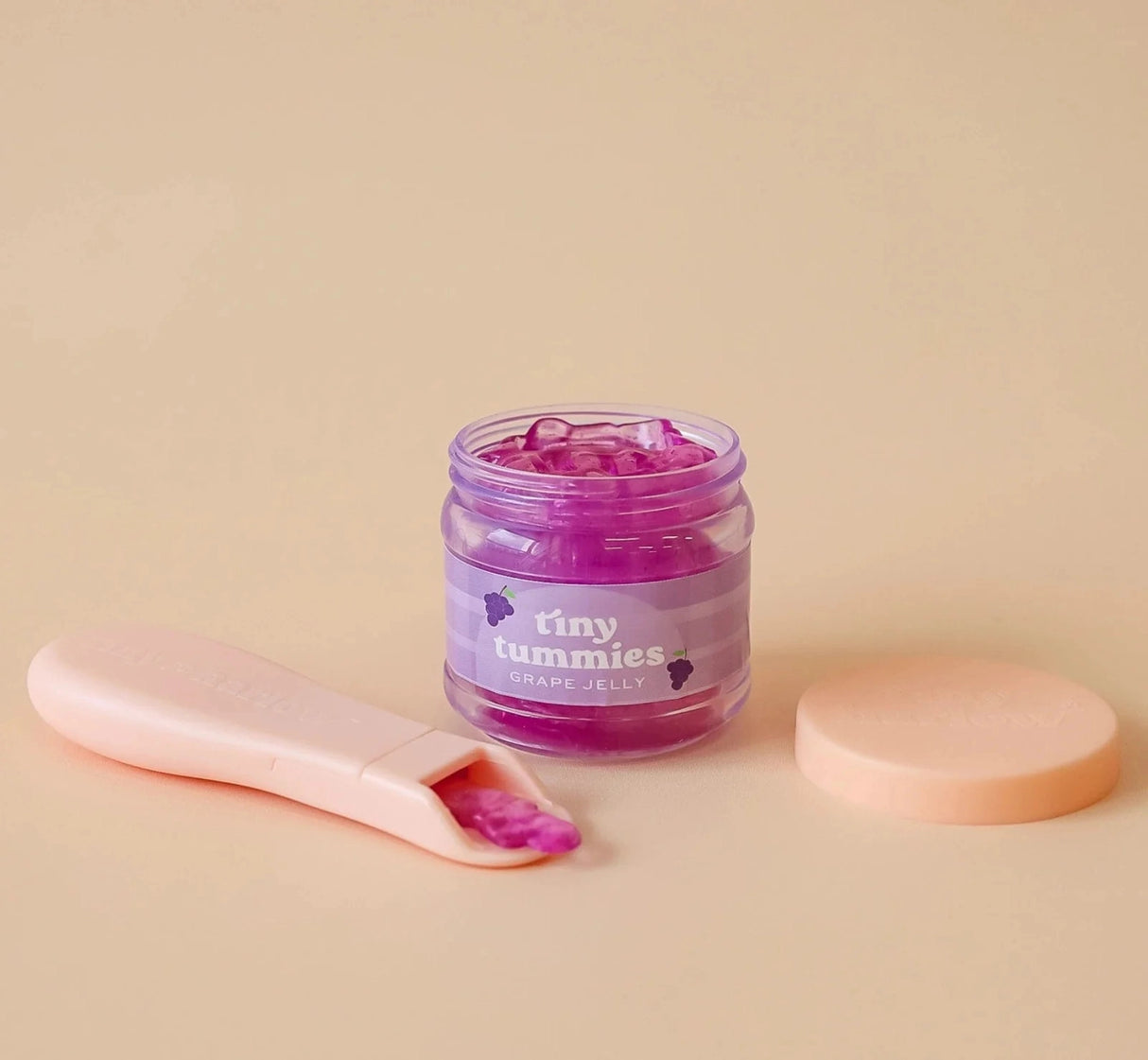 Small jar of grape jelly food with a pastel pink spoon and lid on a soft beige background for dolls.