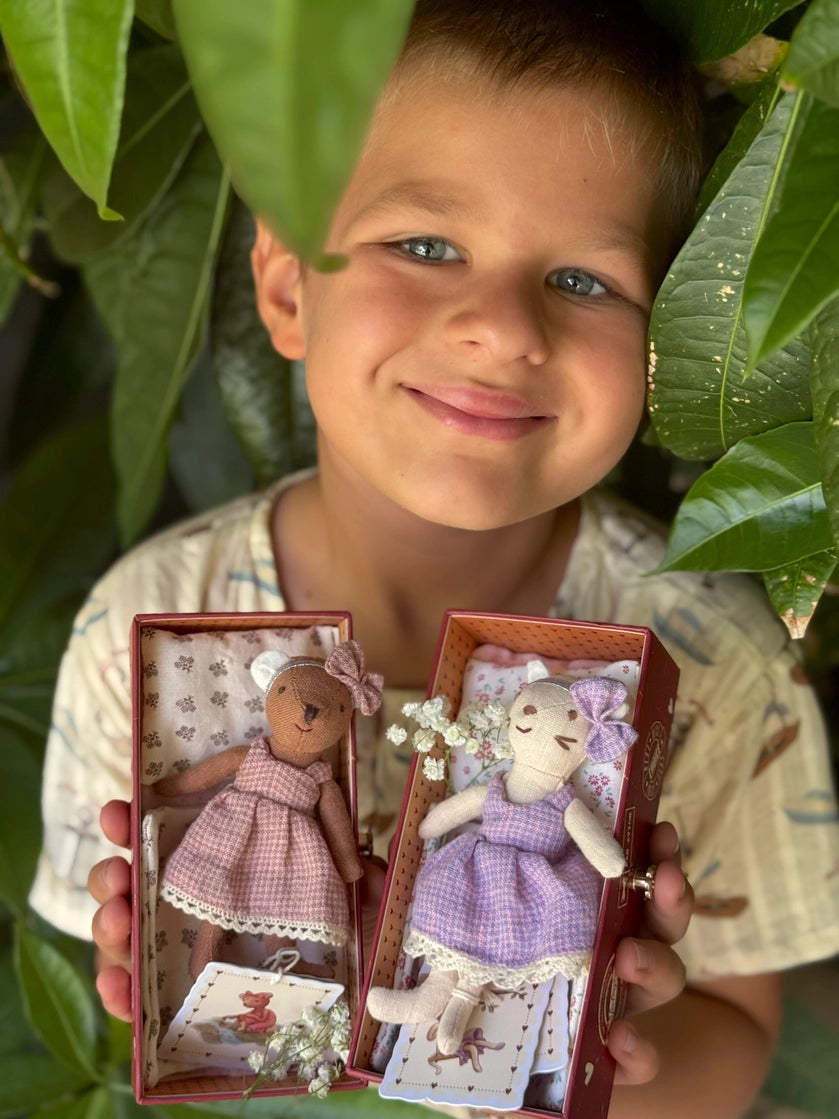Child smiling while holding two fabric cat dolls dressed in purple with lace trim in decorative boxes