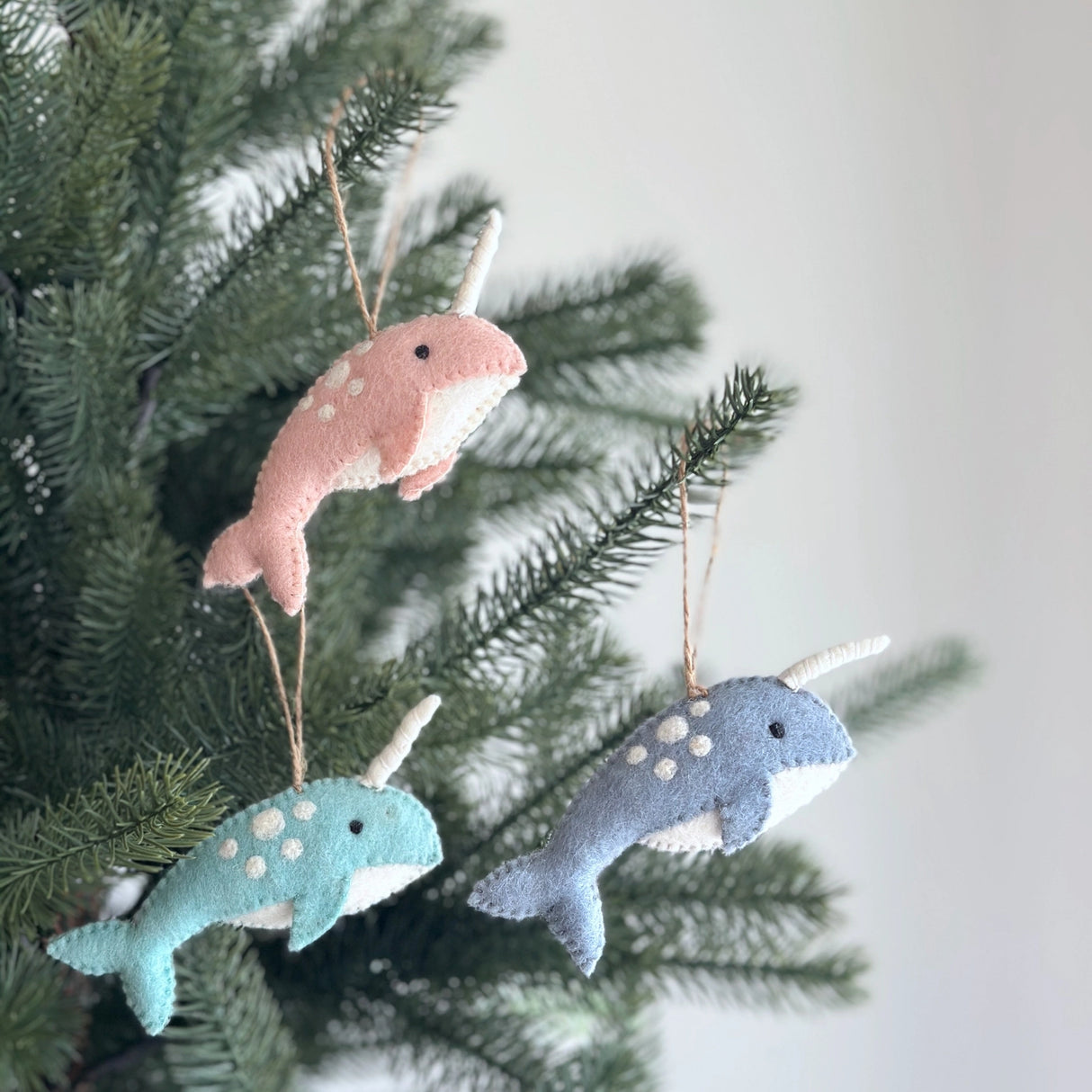 Handmade felt narwhal Christmas ornaments in pastel colors hanging on green pine tree branches