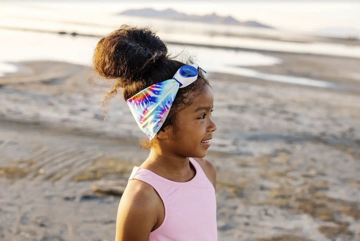 Young girl wearing Goggles Tie Dye with a colorful strap and white frame standing on a sandy beach.