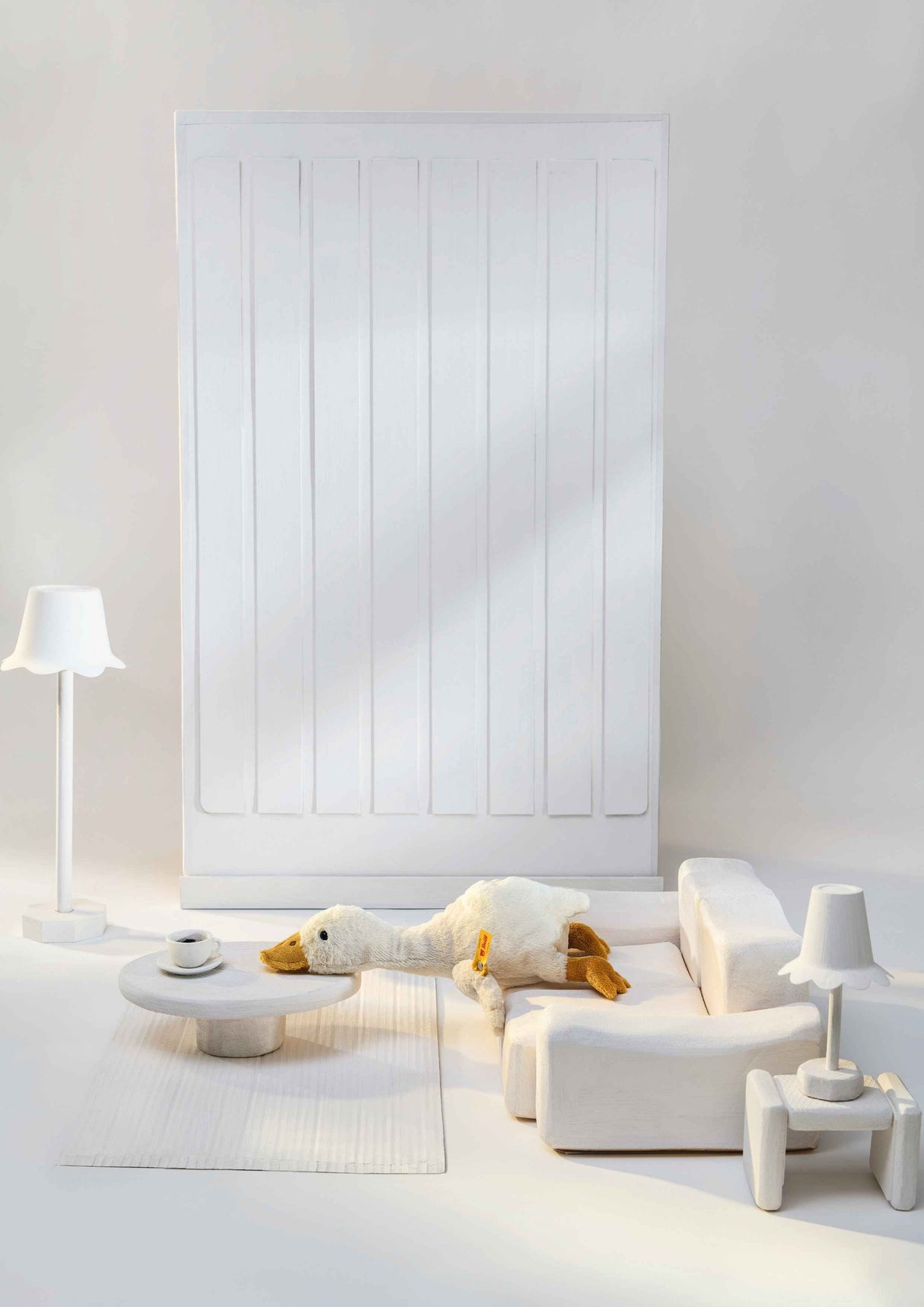 Plush Goose Gilda with long neck and yellow beak resting on a miniature white sofa in a cozy dollhouse setting.