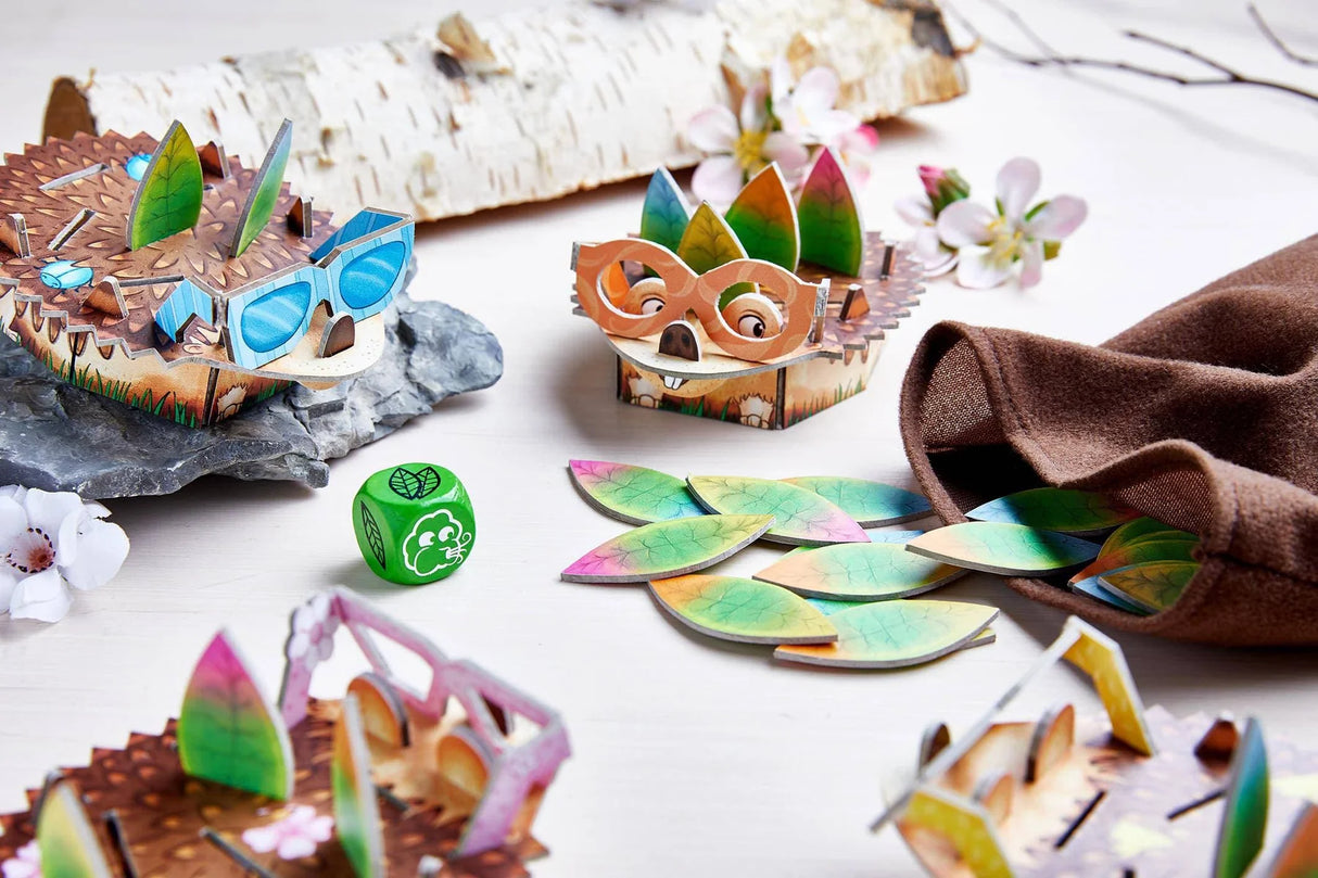 Colorful wooden hedgehog figures with leaves, dice, and cloth bag in a bright, playful memory game setup.