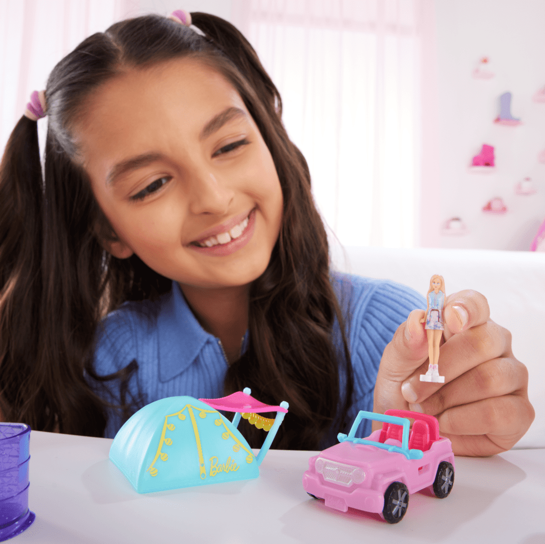 Girl playing with Mini BarbieLand Camping set featuring a pink vehicle, blue tent, and Barbie doll for imaginative play.