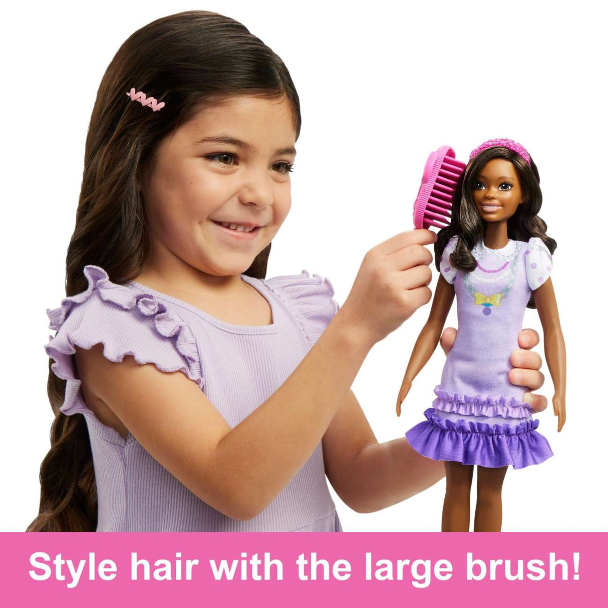A smiling girl styling the hair of My First Barbie Black Hair doll wearing a lilac dress with puff sleeves and a headband.
