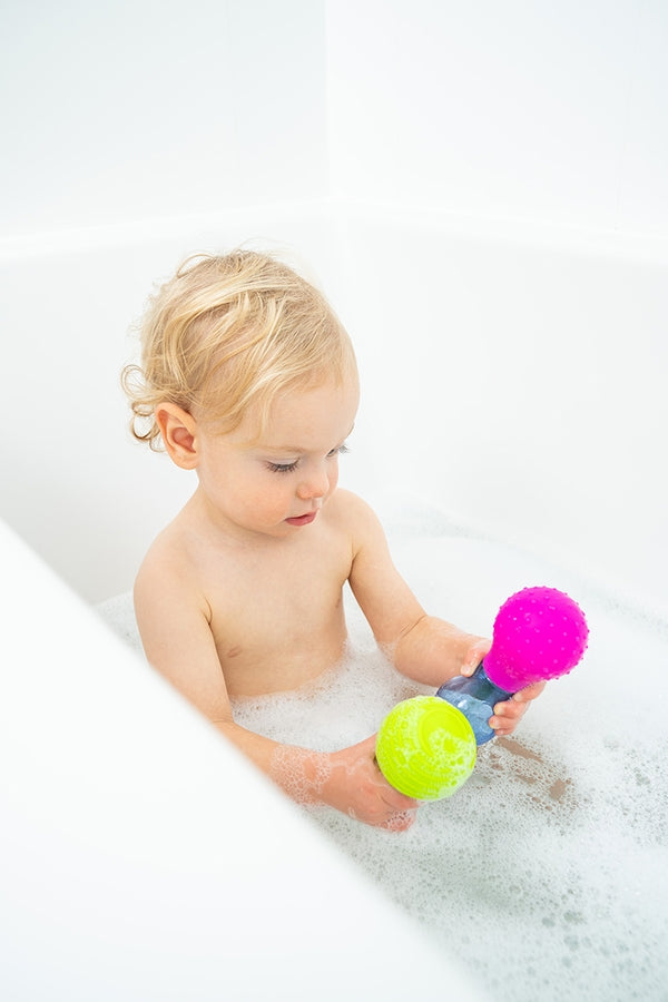 Child playing with colorful Skwooshlz Water Squeeze Bulbs bath toys in a white bathtub filled with bubbles.