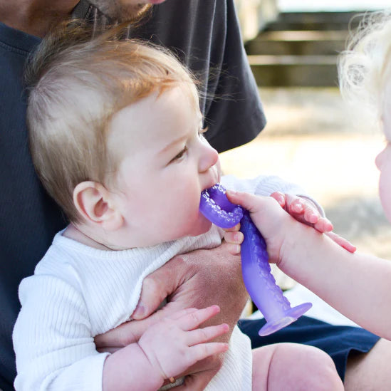 Baby chewing on a purple Tentacle Teether held by another child, soothing sore gums safely and gently.
