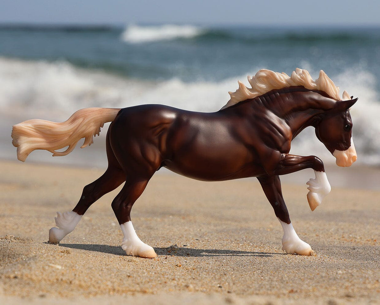 Dark brown model horse with flowing blonde mane and tail walking on a sandy beach with waves in the background