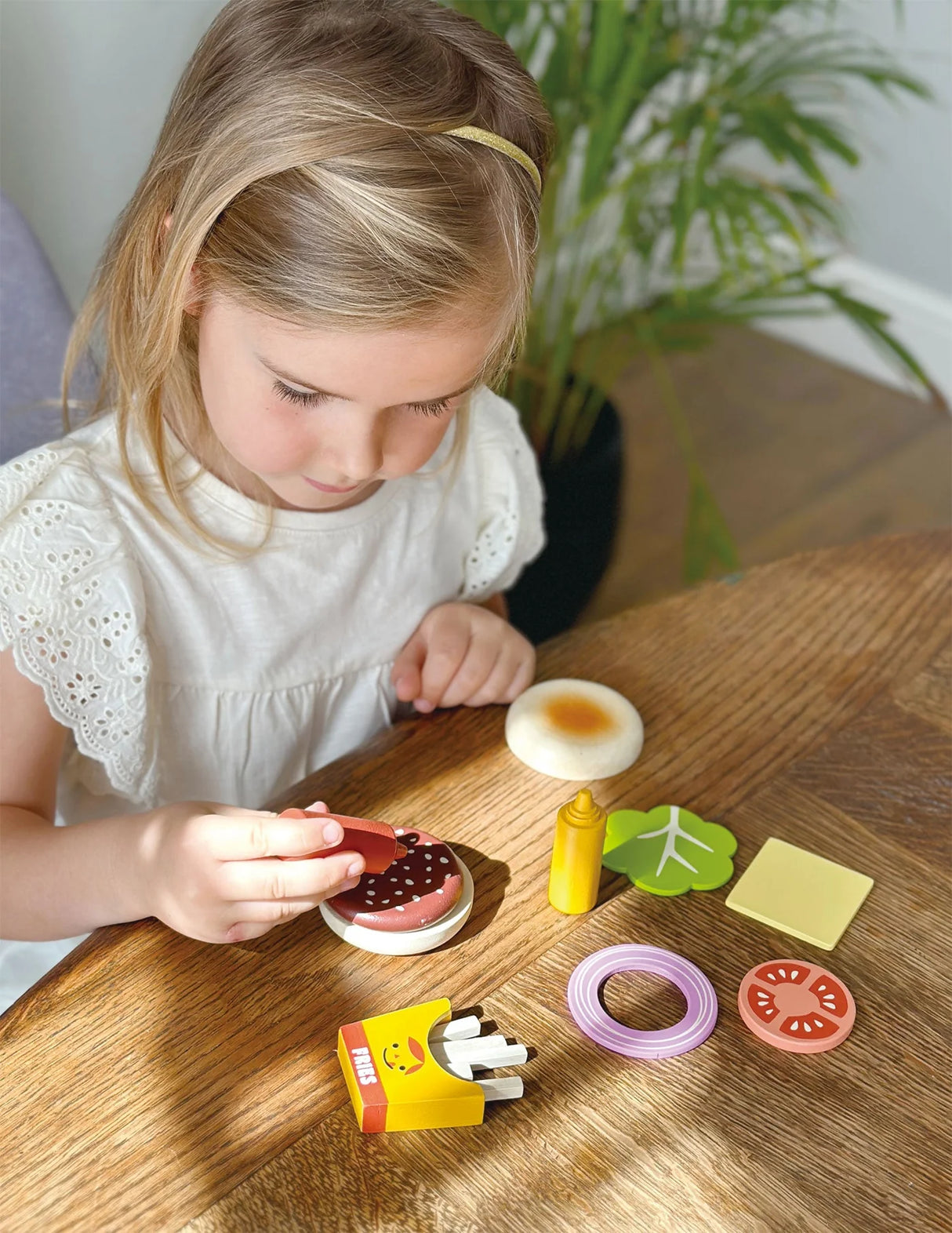 Young girl playing with a wooden take-out burger set including bun, meat patty, cheese, tomato, onion, lettuce, and fries on table