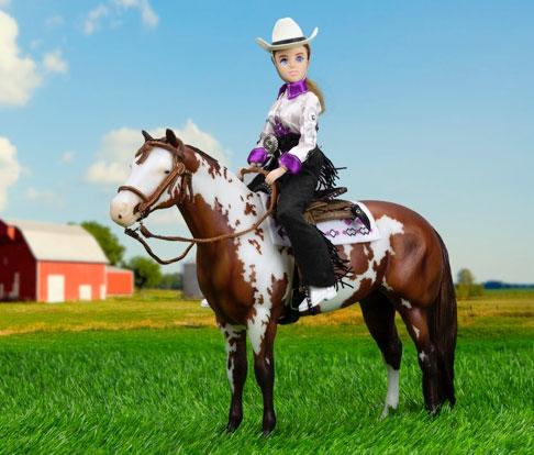 Taylor Cowgirl doll in Western show shirt, hat, and fringed chaps riding a spotted horse on green grass.