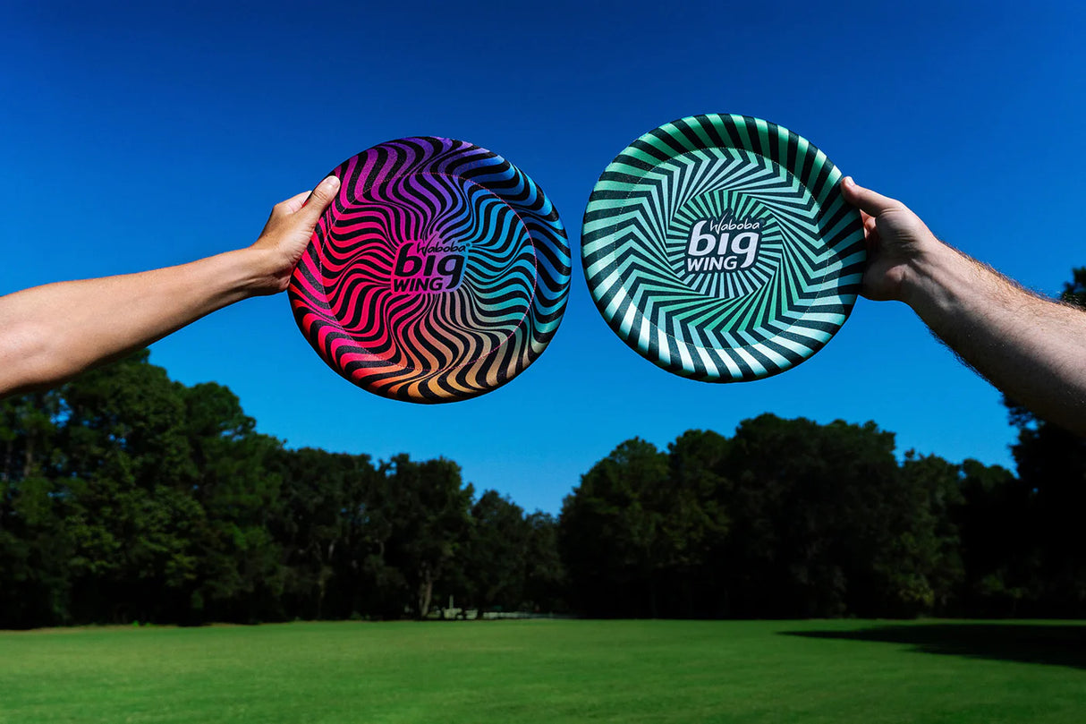 Two hands holding colorful Waboba Big Wing fabric covered discs outdoors on a sunny day.