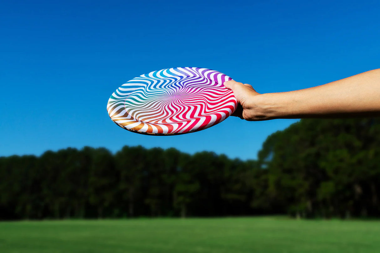 Hand holding a colorful fabric-covered Waboba Big Wing disc against a clear blue sky outdoors.