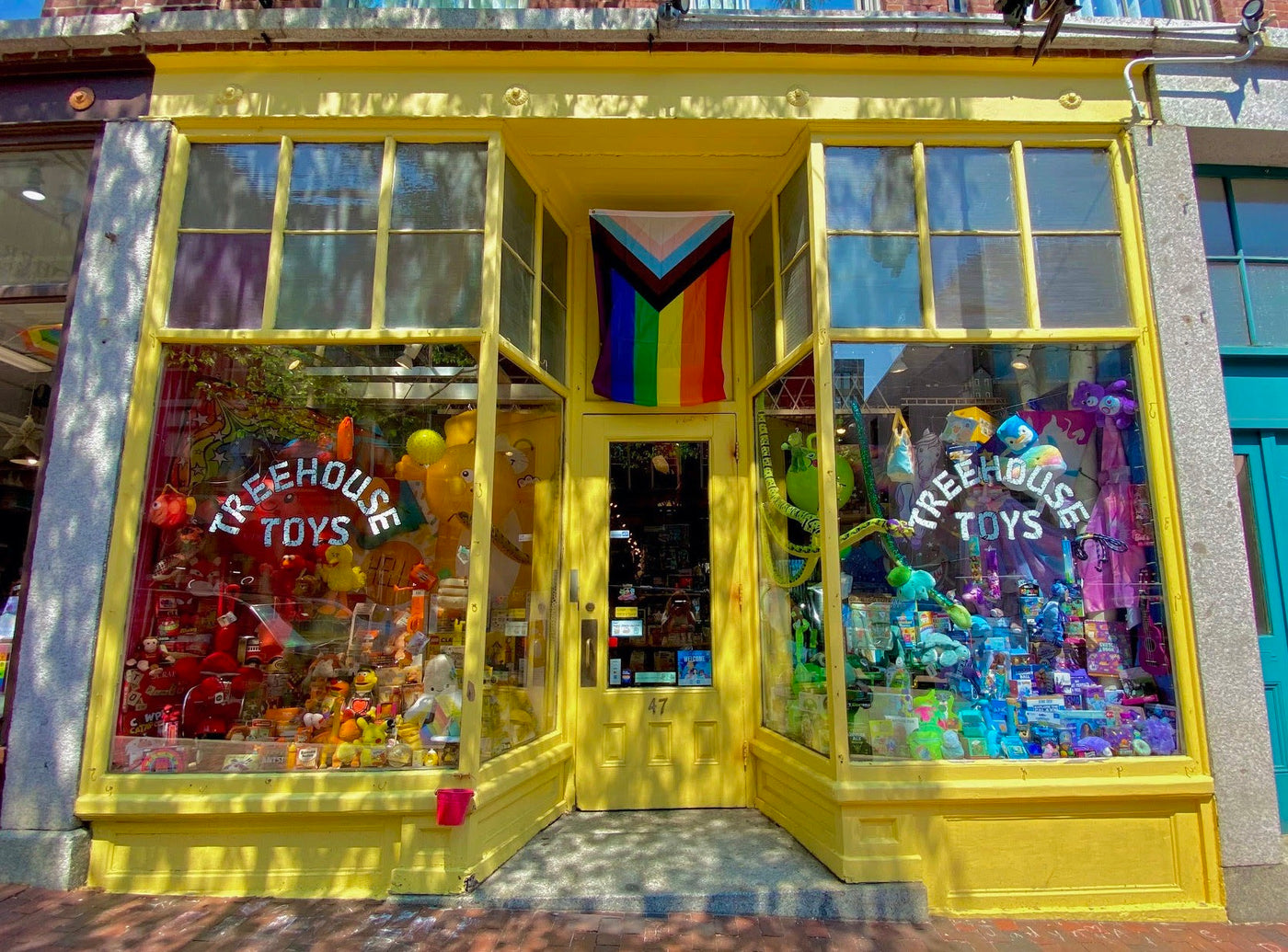 Bright yellow toy store storefront with colorful toys displayed in windows and a pride flag above the entrance