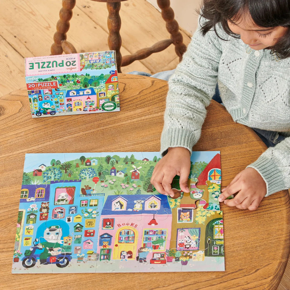 Child completing colorful 20-piece puzzle depicting a lively village with animals, houses, and greenery on wooden table