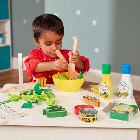Child playing with the Slice & Toss Salad pretend play set, mixing wooden vegetables and felt greens in a yellow bowl.