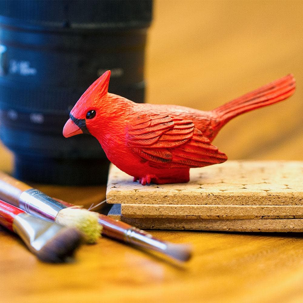 Cardinal figurine brightly hand-painted red with black face details, perched on corkboard beside paintbrushes, product Cardinal.