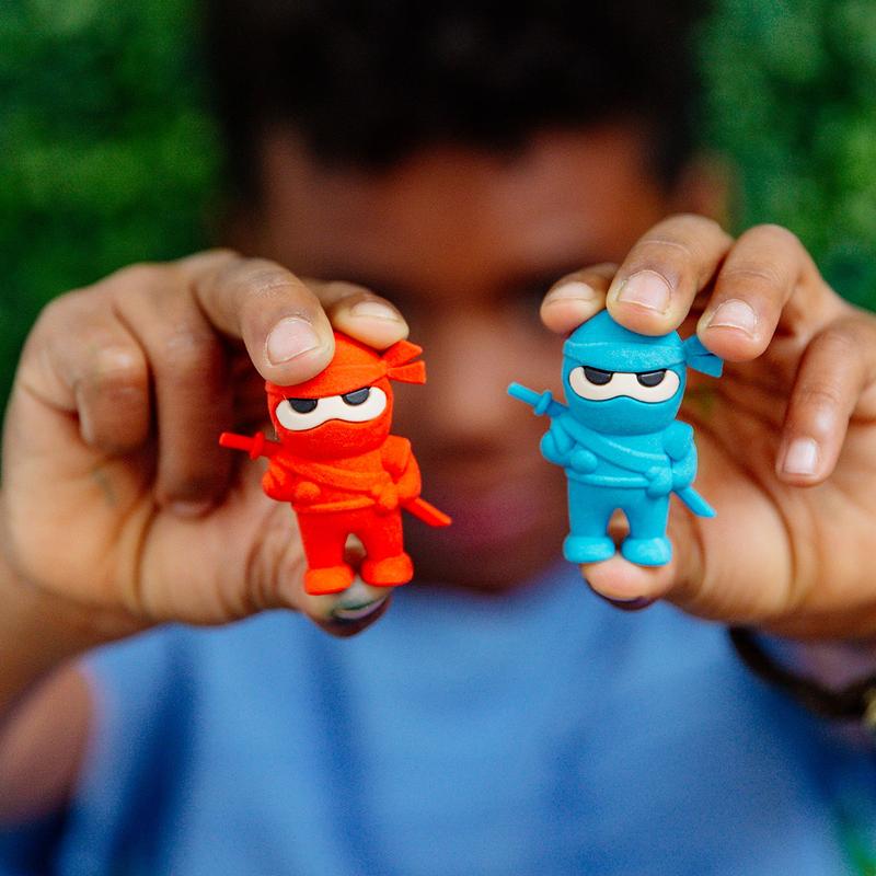 A person holding two Ninja Erasers in red and blue, erasers shaped like ninjas for erasing mistakes.