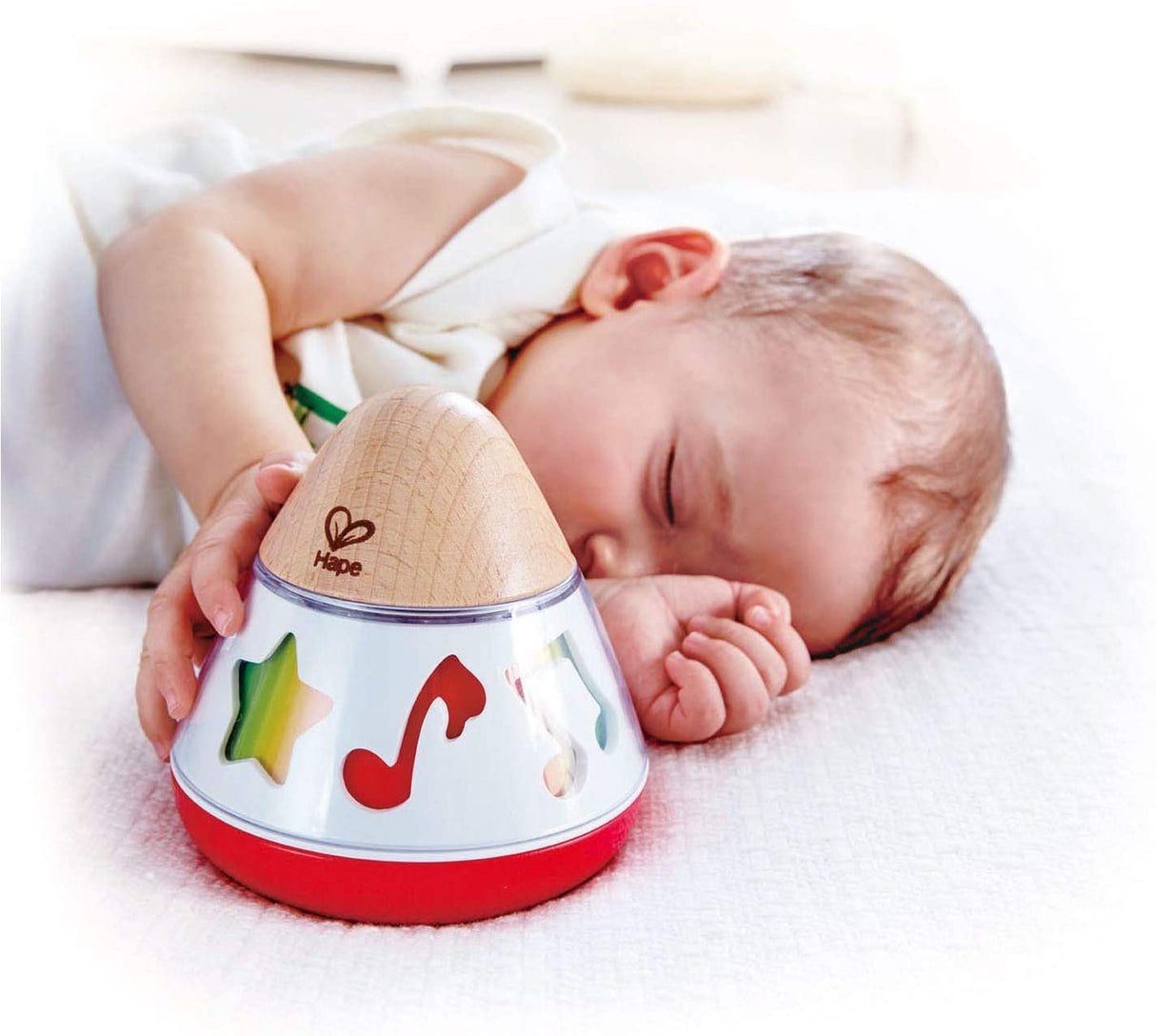 Sleeping baby holding the Music Box with colorful musical symbols and a wooden top on a soft white surface.