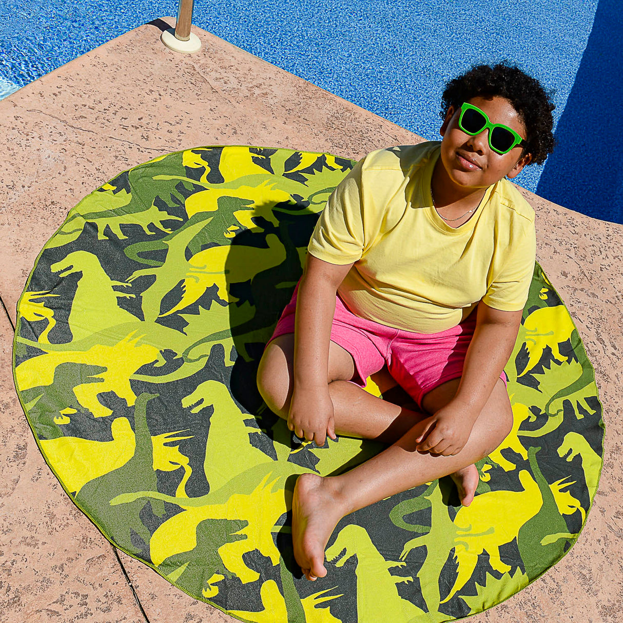 Child sitting on a vibrant Towel Dino Camo round towel by the pool, perfect for beach or picnic use.