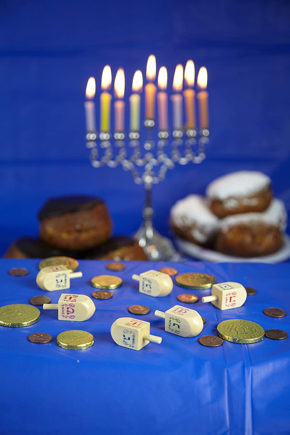 Wooden Dreidel with traditional Hebrew letters on blue table surrounded by Hanukkah gelt and a lit menorah in background