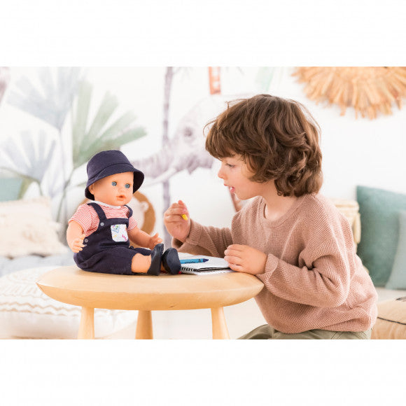 Child playing with Grand Poupon Augustin doll dressed in navy outfit and sun hat seated on a small wooden table.