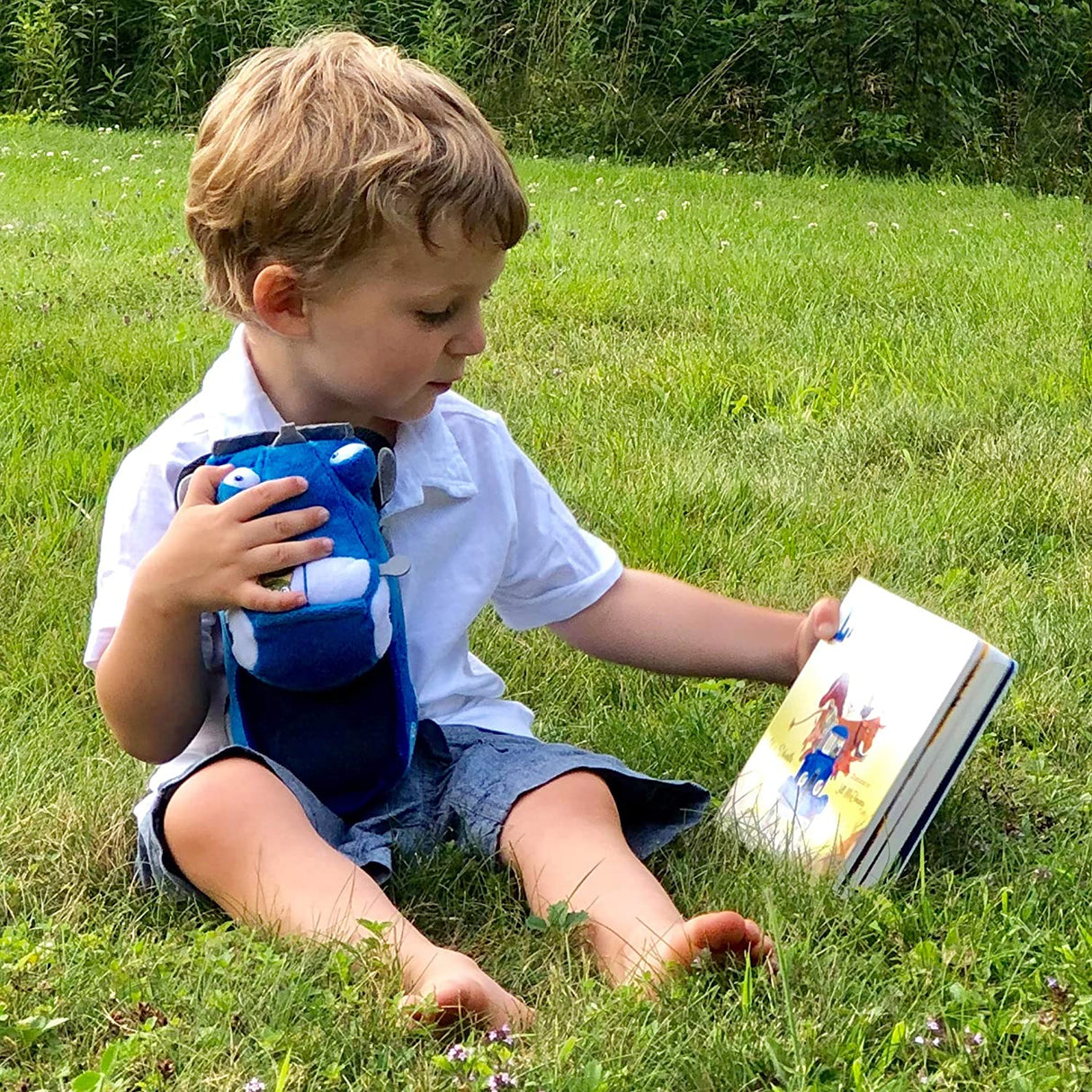 Child sitting on grass holding the Little Blue Truck soft toy and reading a book outdoors.