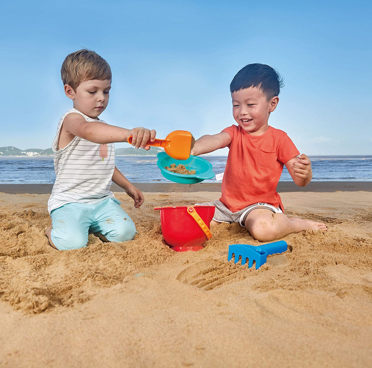 Two children playing with the Beach Basics 4pc Set including bucket, shovel, sieve, and rake on the beach sand.