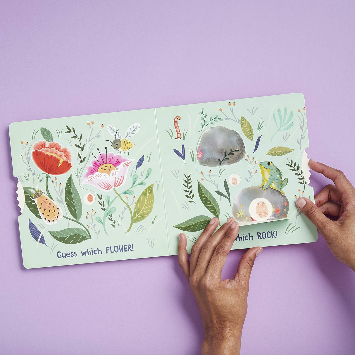 Hands holding the interactive toddler board book Eggs are Everywhere with colorful flowers and rocks illustrations.