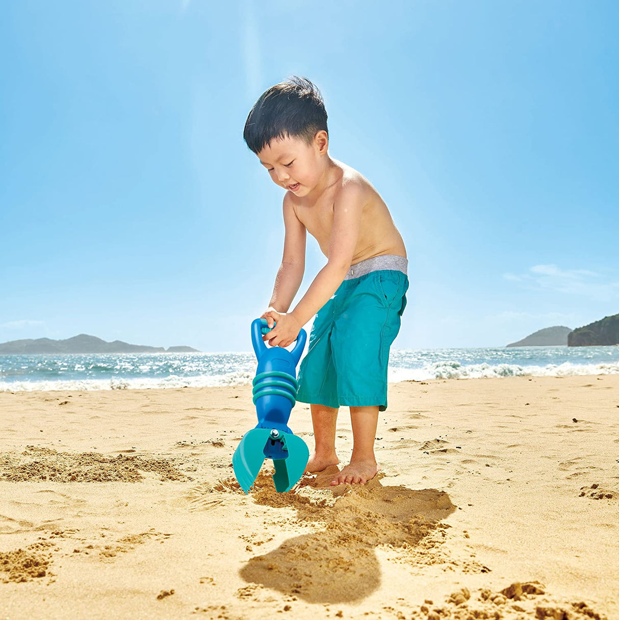 Young boy on beach using the Grabber Blue sand toy to dig and scoop sand under a clear sky.