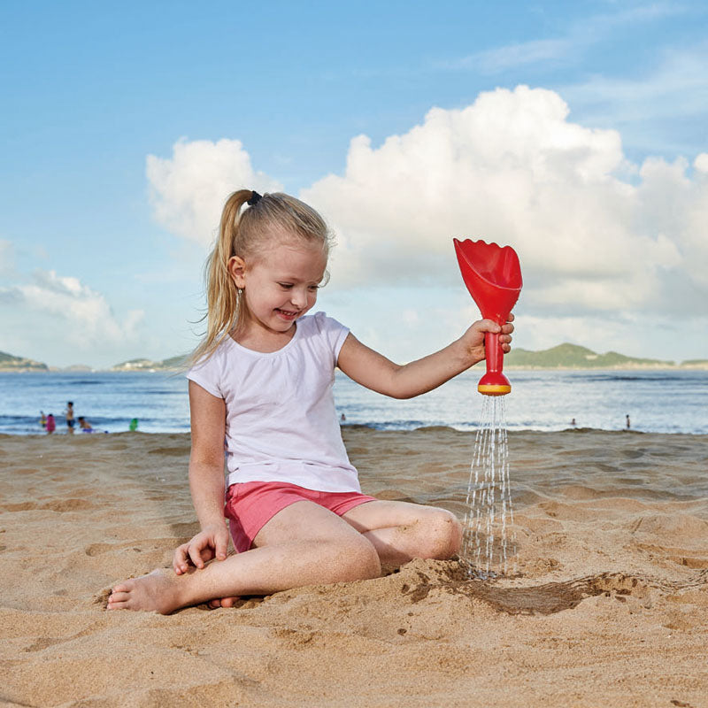 Child playing on the beach using the Rain Shovel Red toy to pour water and sand for fun and exploration.