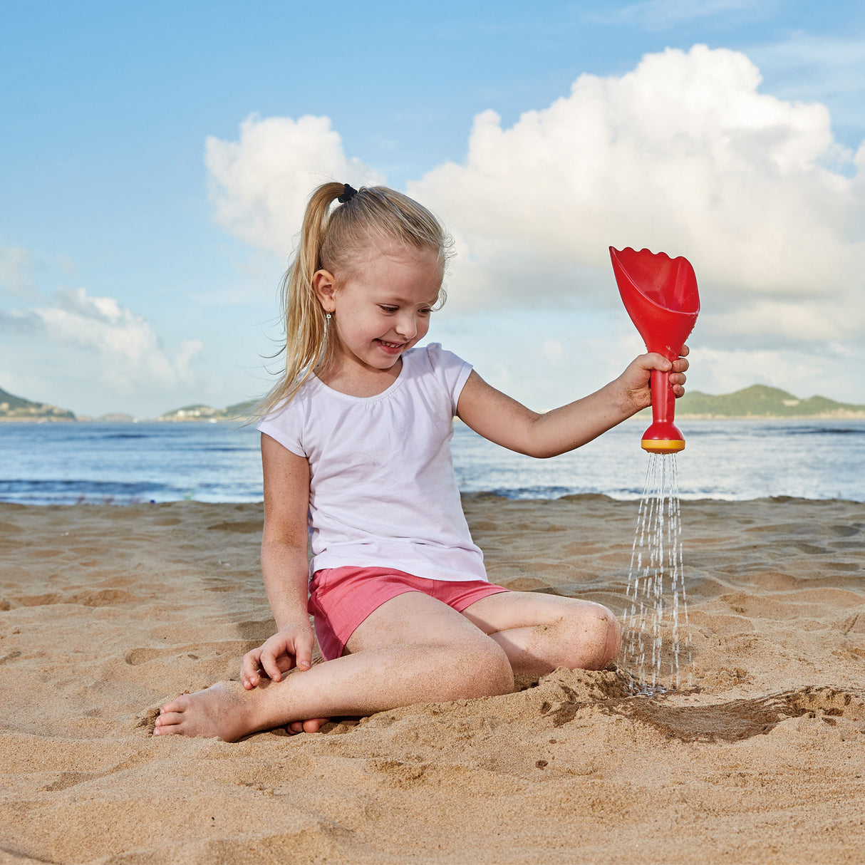 Young girl playing with a red Rain Shovel Blue toy on the sandy beach under a clear sky